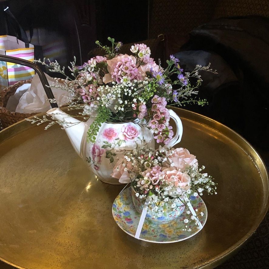 Floral arrangement in a porcelain teapot and teacup, filled with pink and white flowers, set on a gold tray.