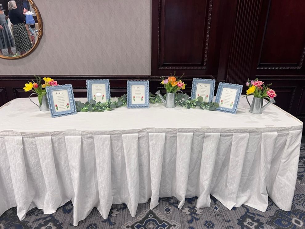 A table with framed cards and floral arrangements. The cards are in light blue frames and sit on a white tablecloth.