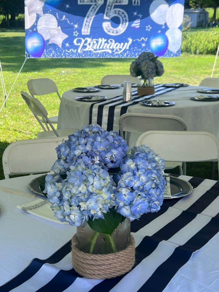 Blue hydrangea flowers in a glass jar with rope detail, centerpieces on a table set for a 75th birthday party with blue and white decor.