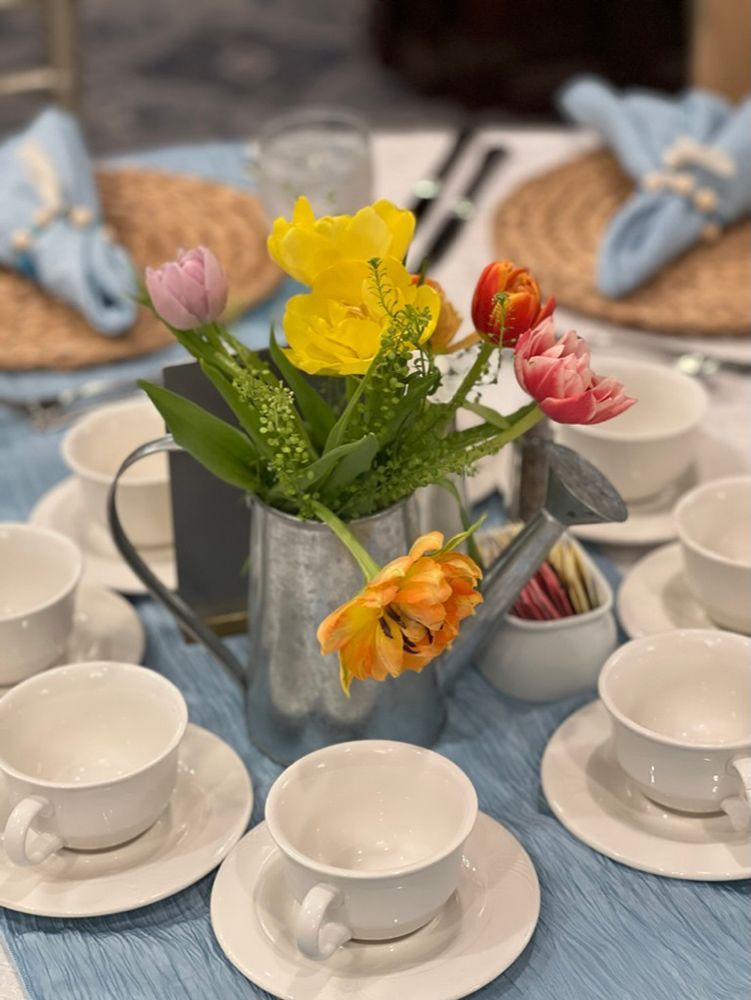 Table setting with teacups, napkins, and a floral centerpiece in a watering can. Tulips in yellow, pink, and orange adorn the table.