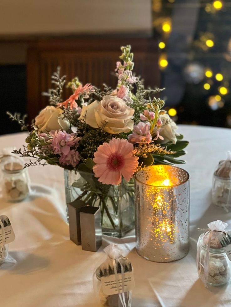 A floral centerpiece with roses and gerberas on a white tablecloth, next to a lit candle and favors.