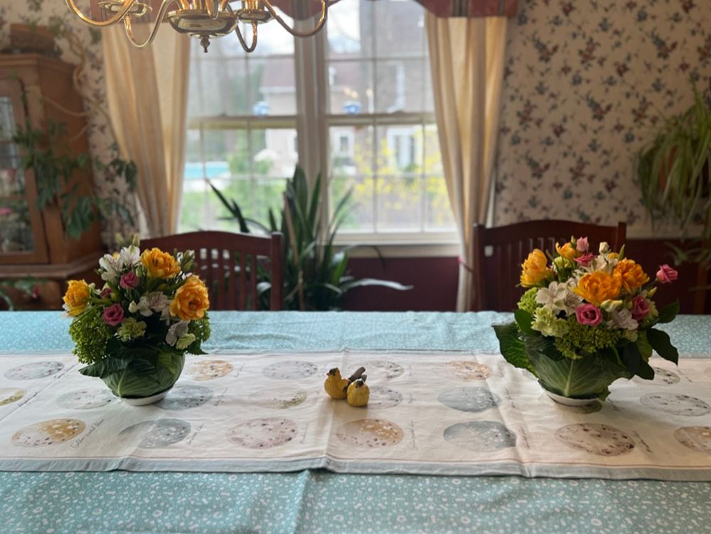 Two floral centerpieces on a table set for a meal; the room has floral wallpaper, a window, and a chandelier.