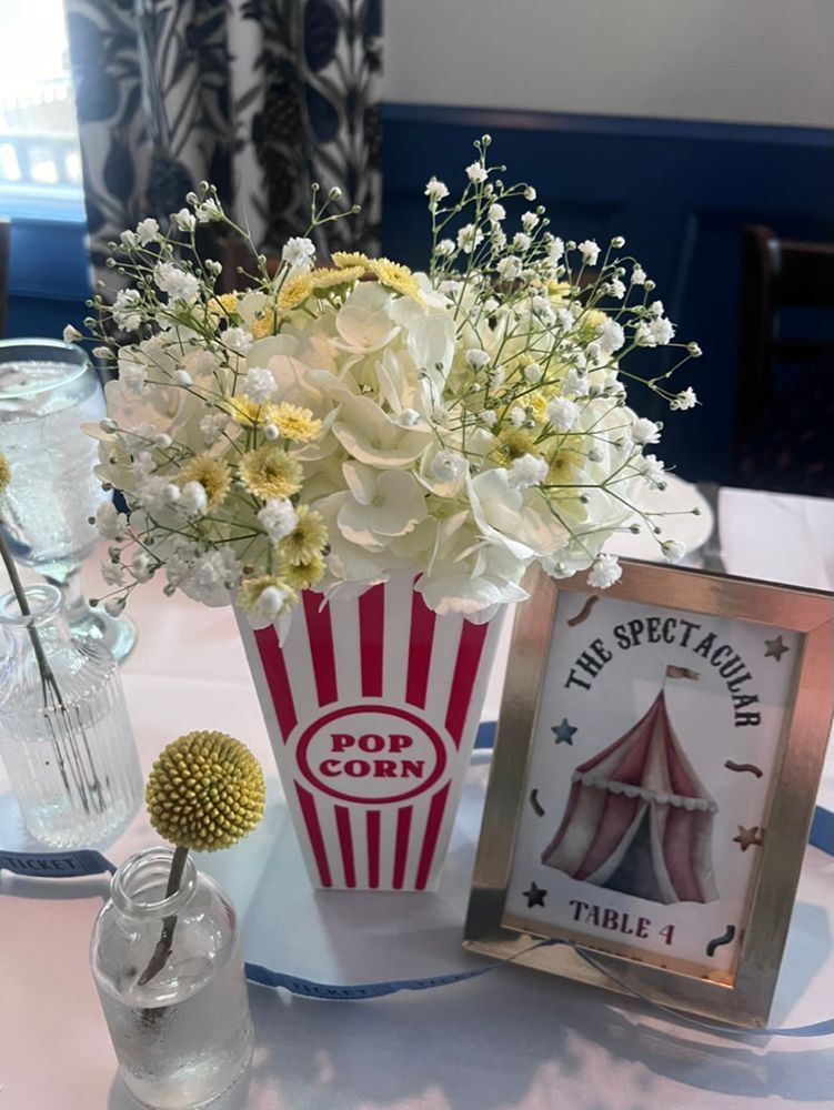 A circus-themed table centerpiece with white and yellow flowers in a popcorn container, and a framed table number.
