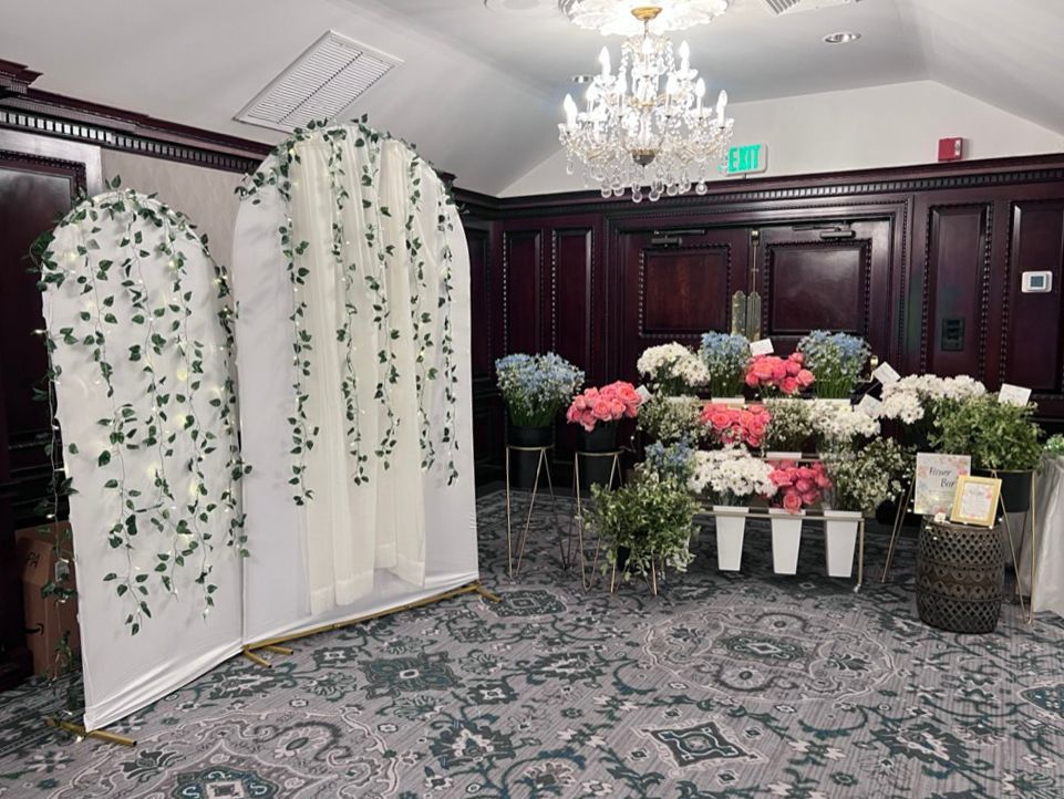 A flower shop display at an event. Features include an archway with faux vines, various flower buckets, and a crystal chandelier in the background.