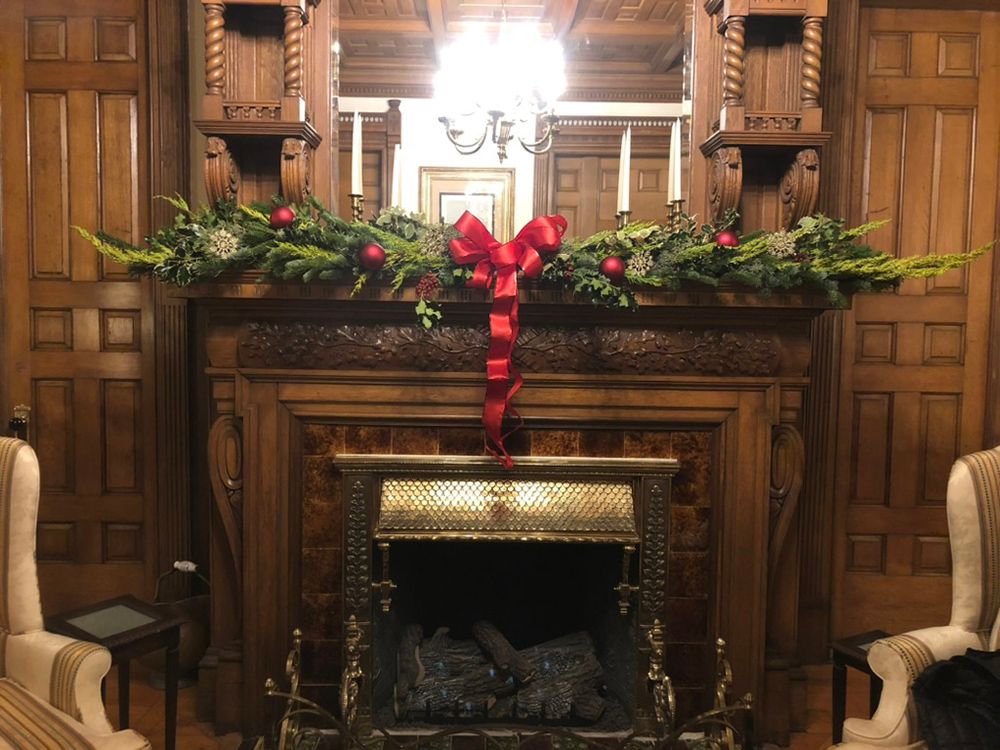 Ornate wooden fireplace decorated with evergreen garland, red bows, and ornaments. The setting is a room with wood-paneled walls.
