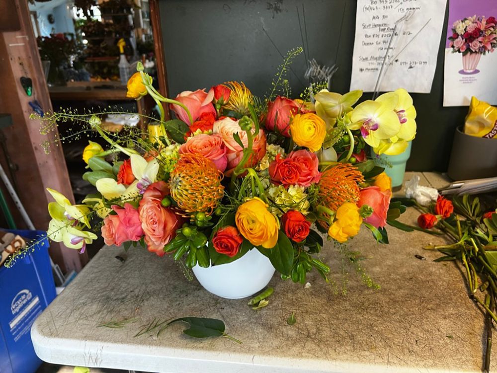 A vibrant floral arrangement in a white bowl on a work surface, featuring orange, yellow, and pink flowers.