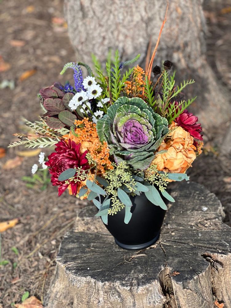 A fall floral arrangement in a black vase sitting on a tree stump. It features a large purple and green cabbage with various colorful blooms and foliage.