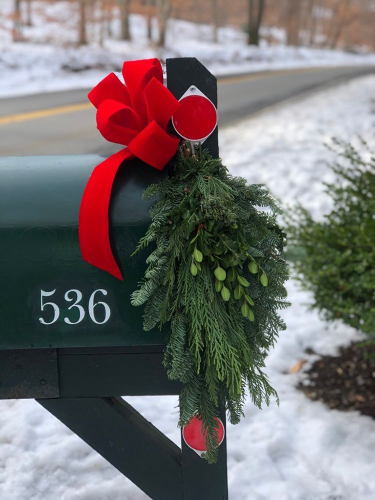A green mailbox decorated for Christmas with a red bow, greenery, and ornaments; snow in the background.