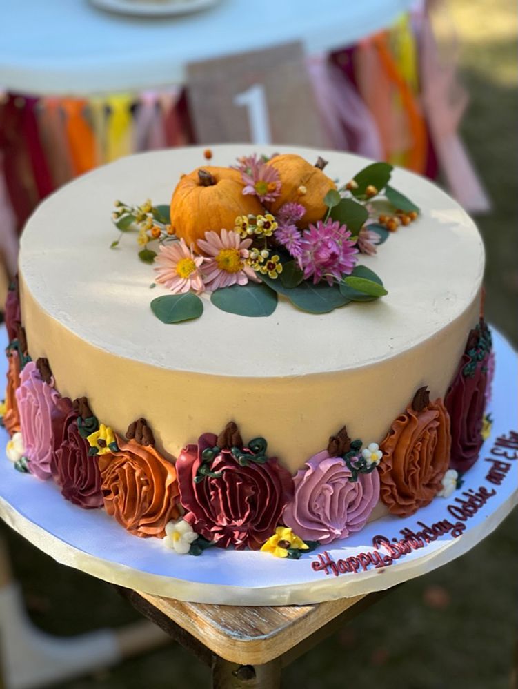 A fall-themed cake with buttercream flowers in autumn colors, decorated with miniature pumpkins and flowers, sits on a table.