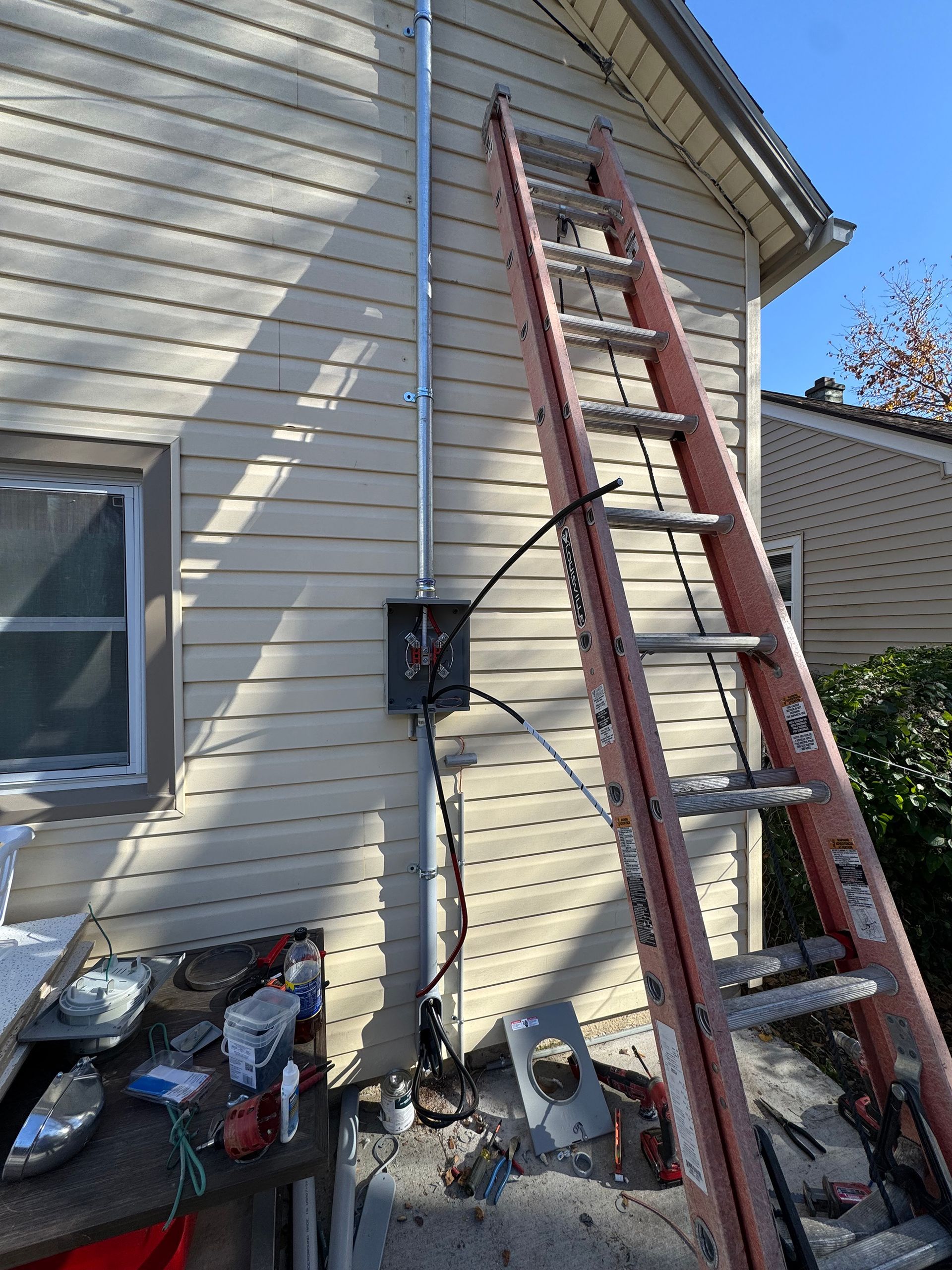 Ladder leaning against a house with electrical work in progress, tools on ground.