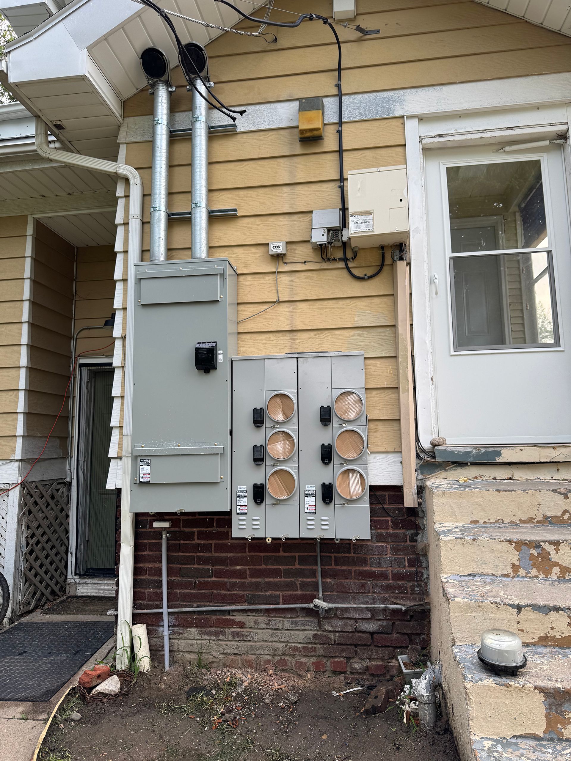 Electrical panel on exterior of a yellow house with multiple meter sockets and conduit.