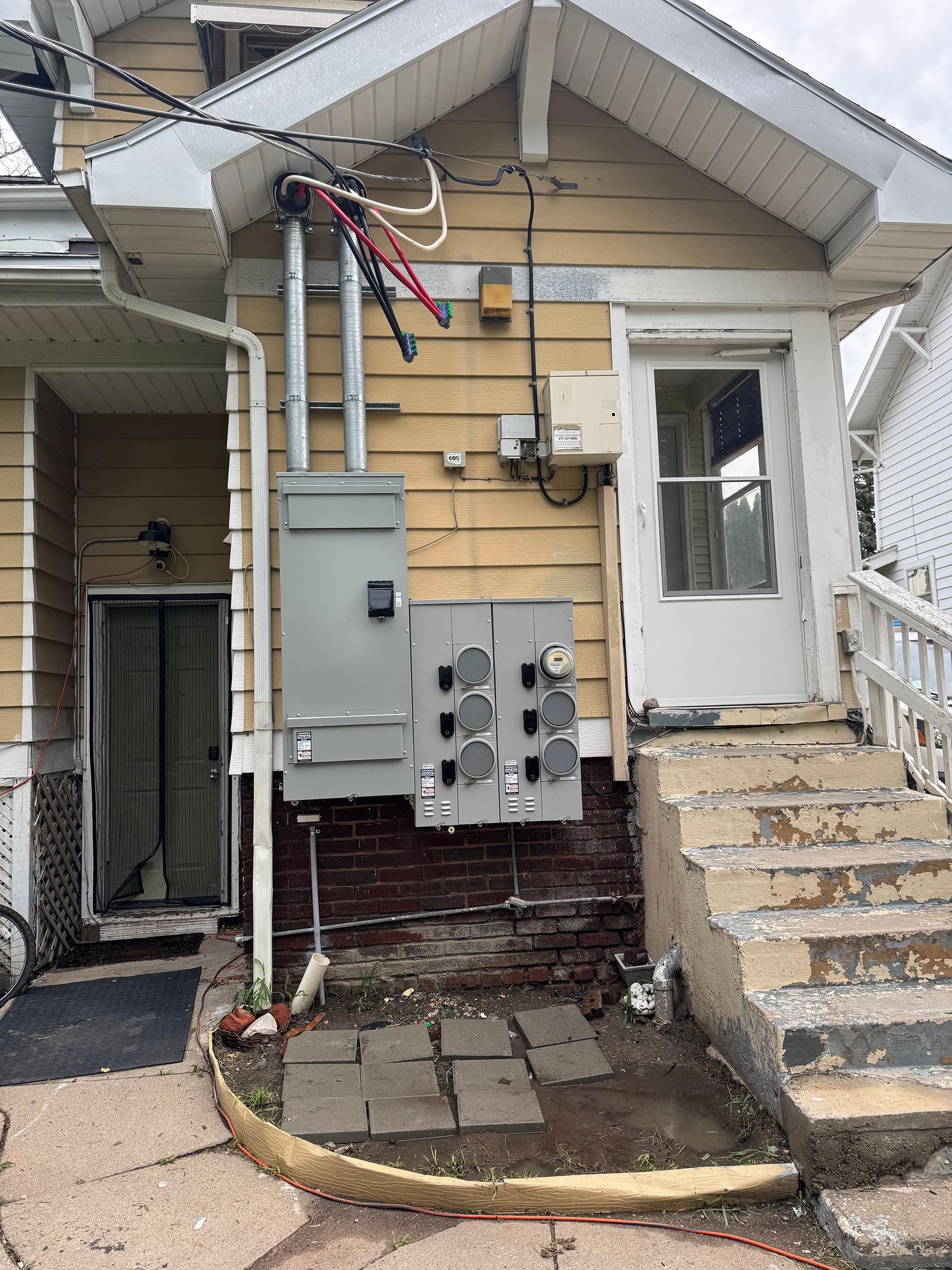 Exterior of a house with electrical panels and a back door. Yellow siding, brick foundation, and concrete steps.
