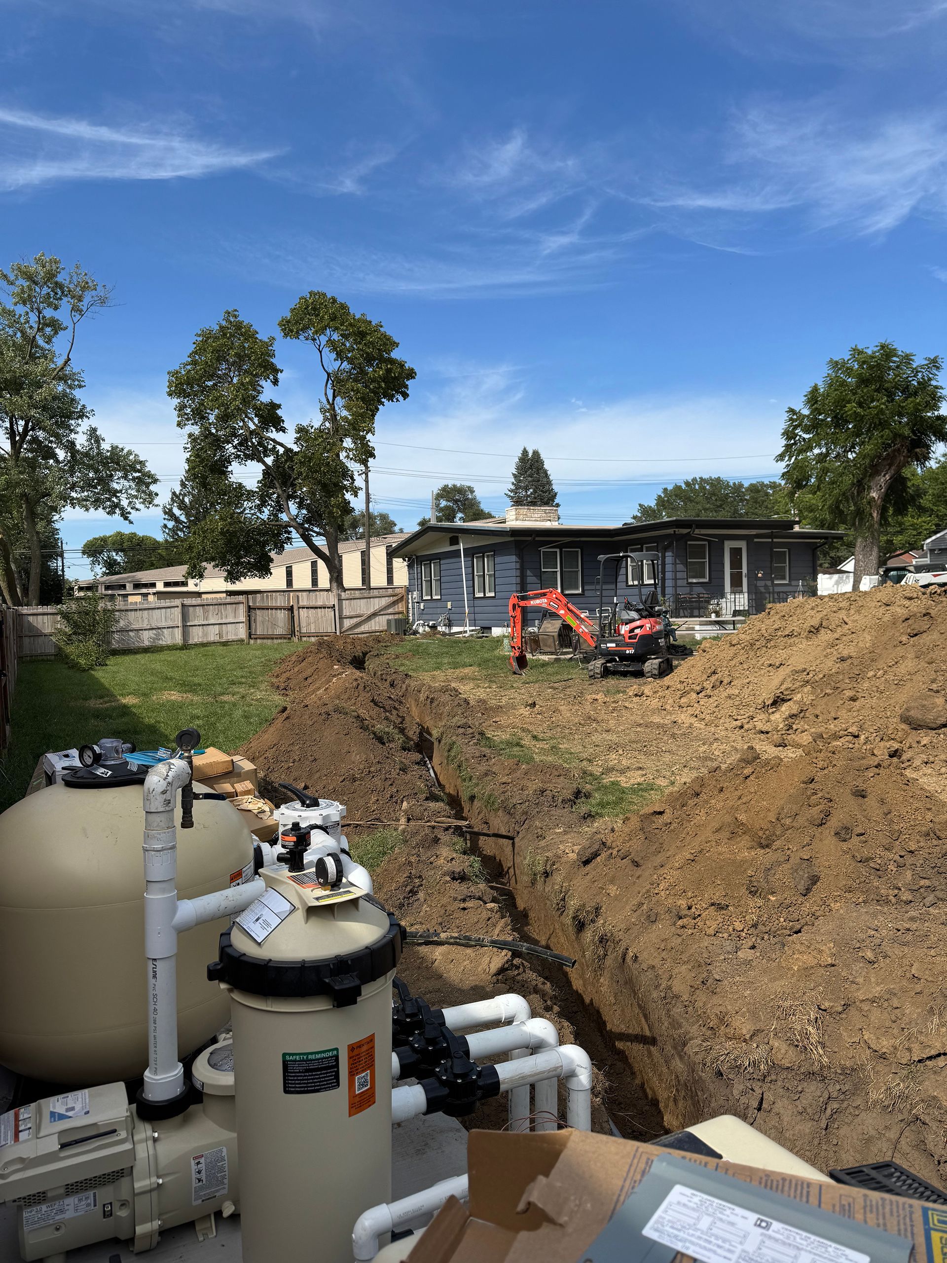 Pool equipment in foreground, trench being dug in backyard with small excavator. Blue sky, house in the background.