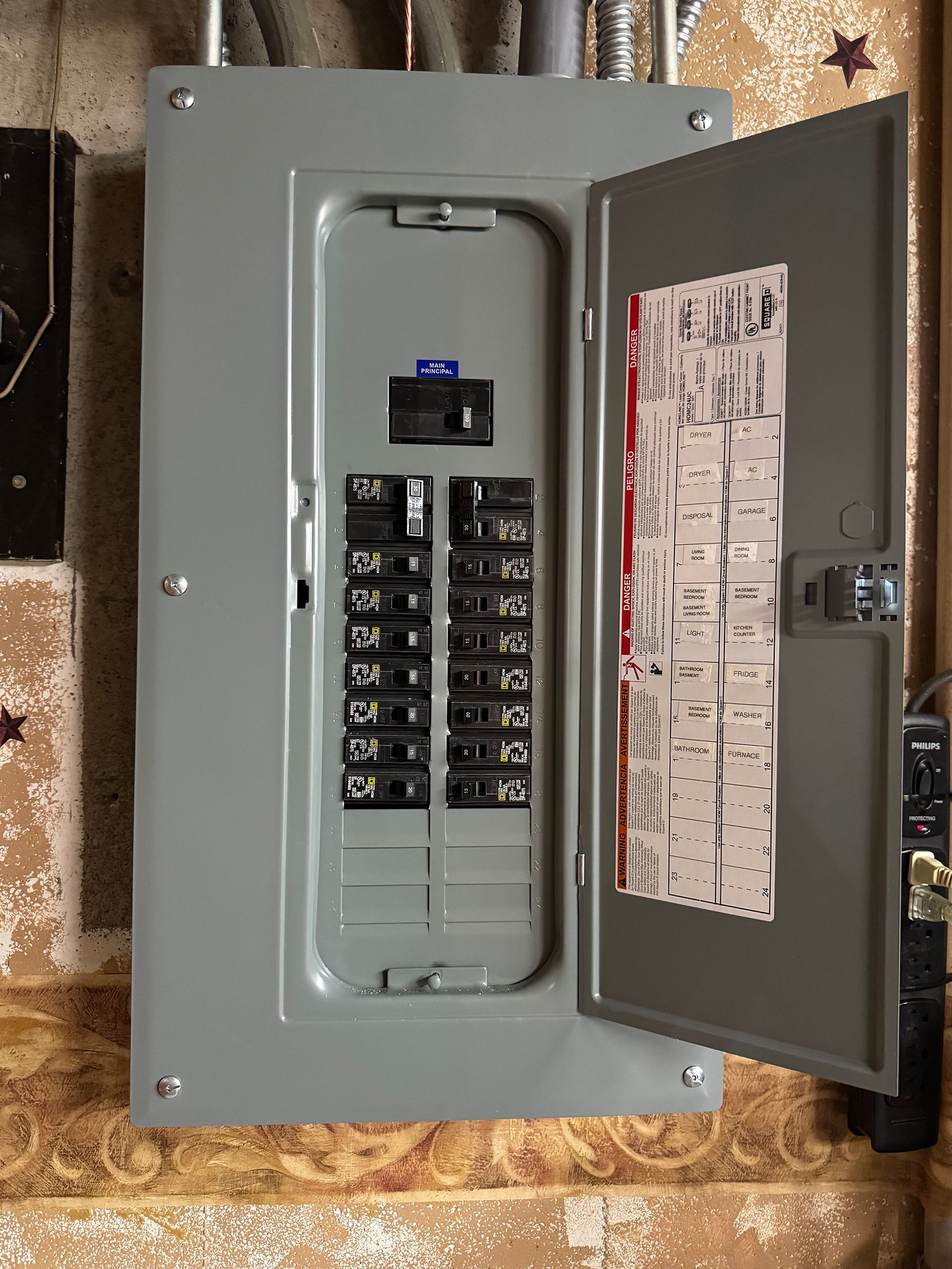 Gray electrical panel with open door, showing circuit breakers. Mounted on a beige wall.