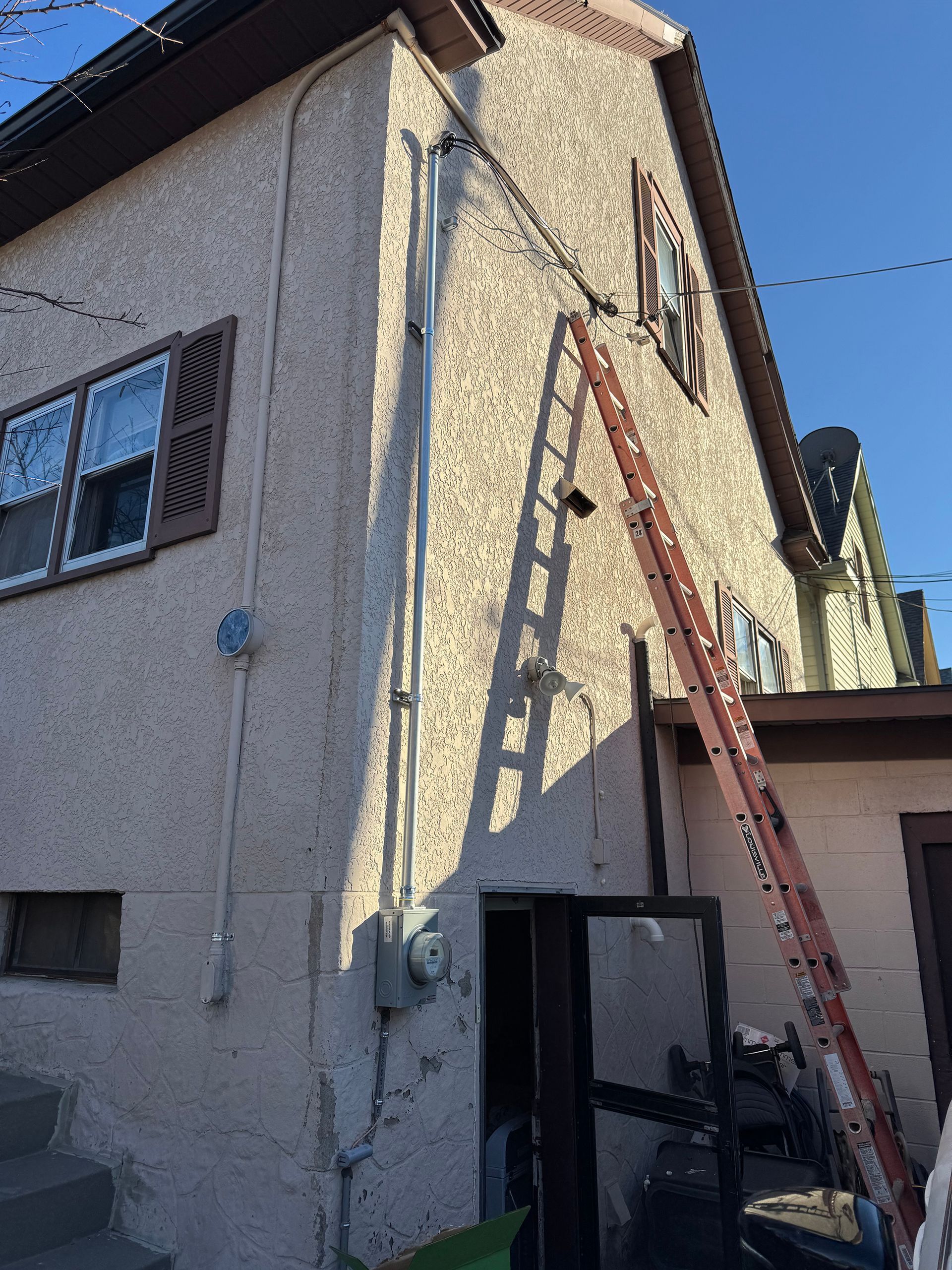 Side of a two-story beige building with a ladder leaning against it.  Electrical conduit runs up the wall. Open door.