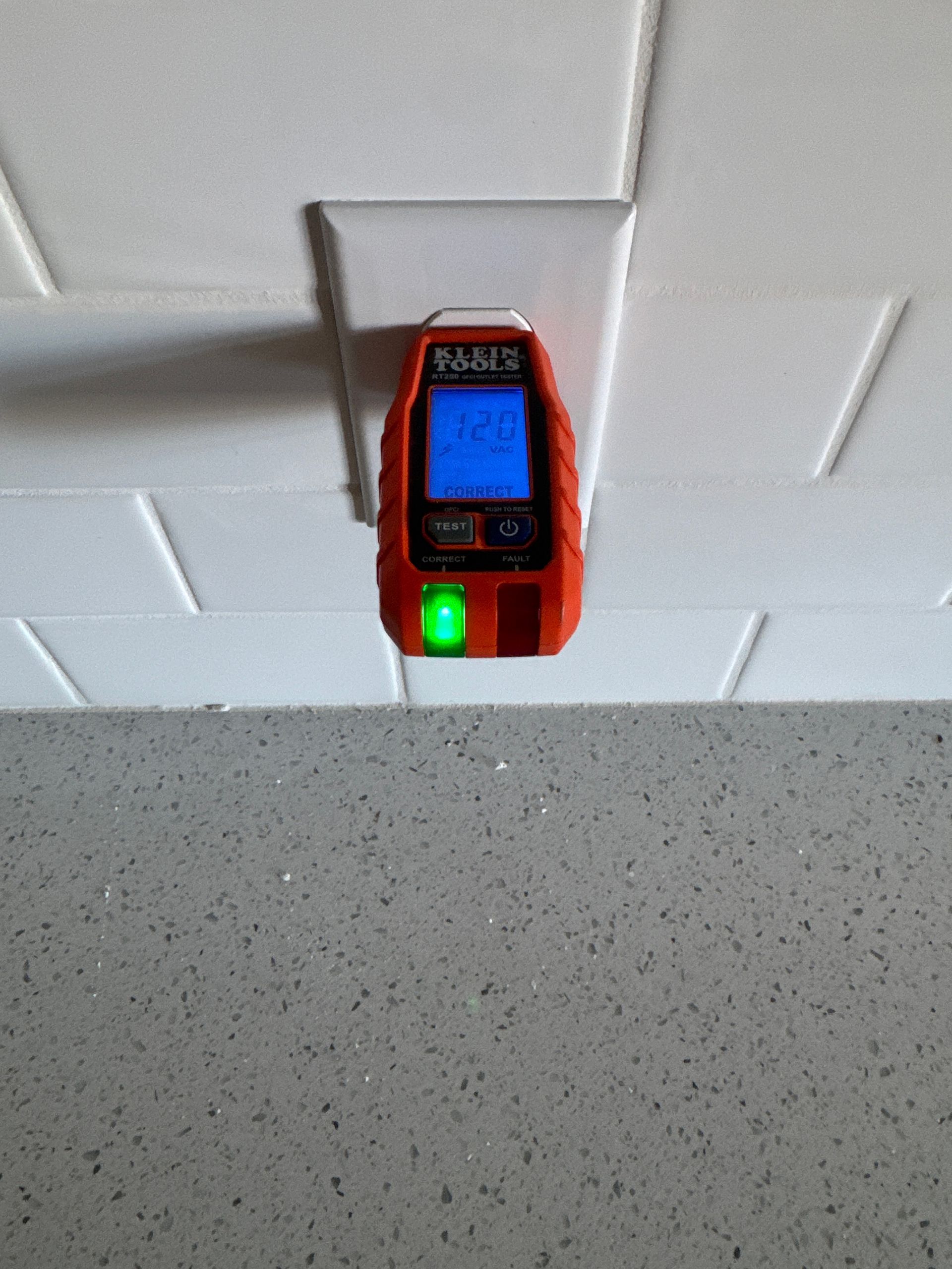 Orange stud finder detecting behind a white outlet in front of a gray countertop.