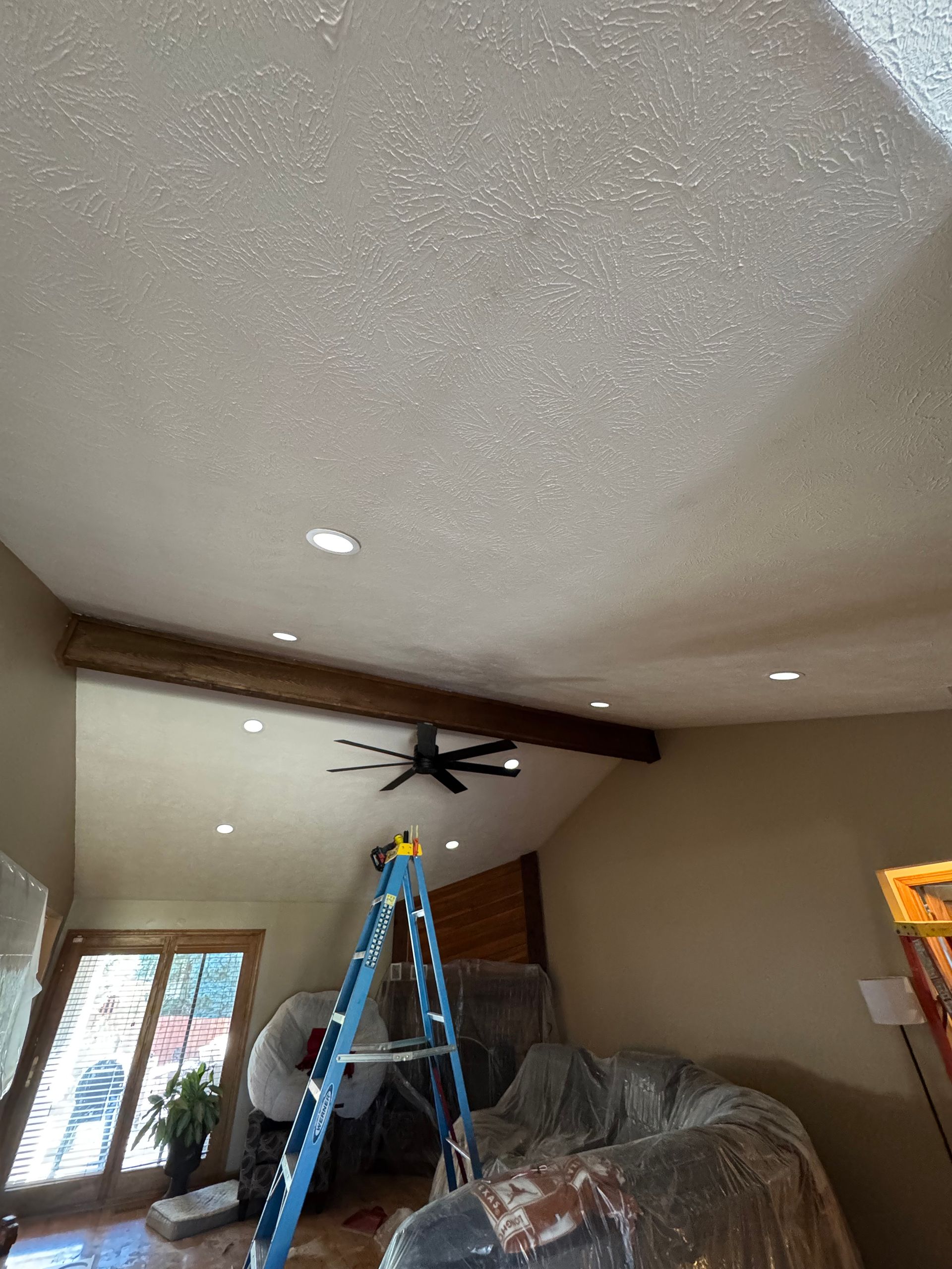 A room with a textured ceiling, recessed lights, and a wooden beam. A ladder stands near a black ceiling fan.