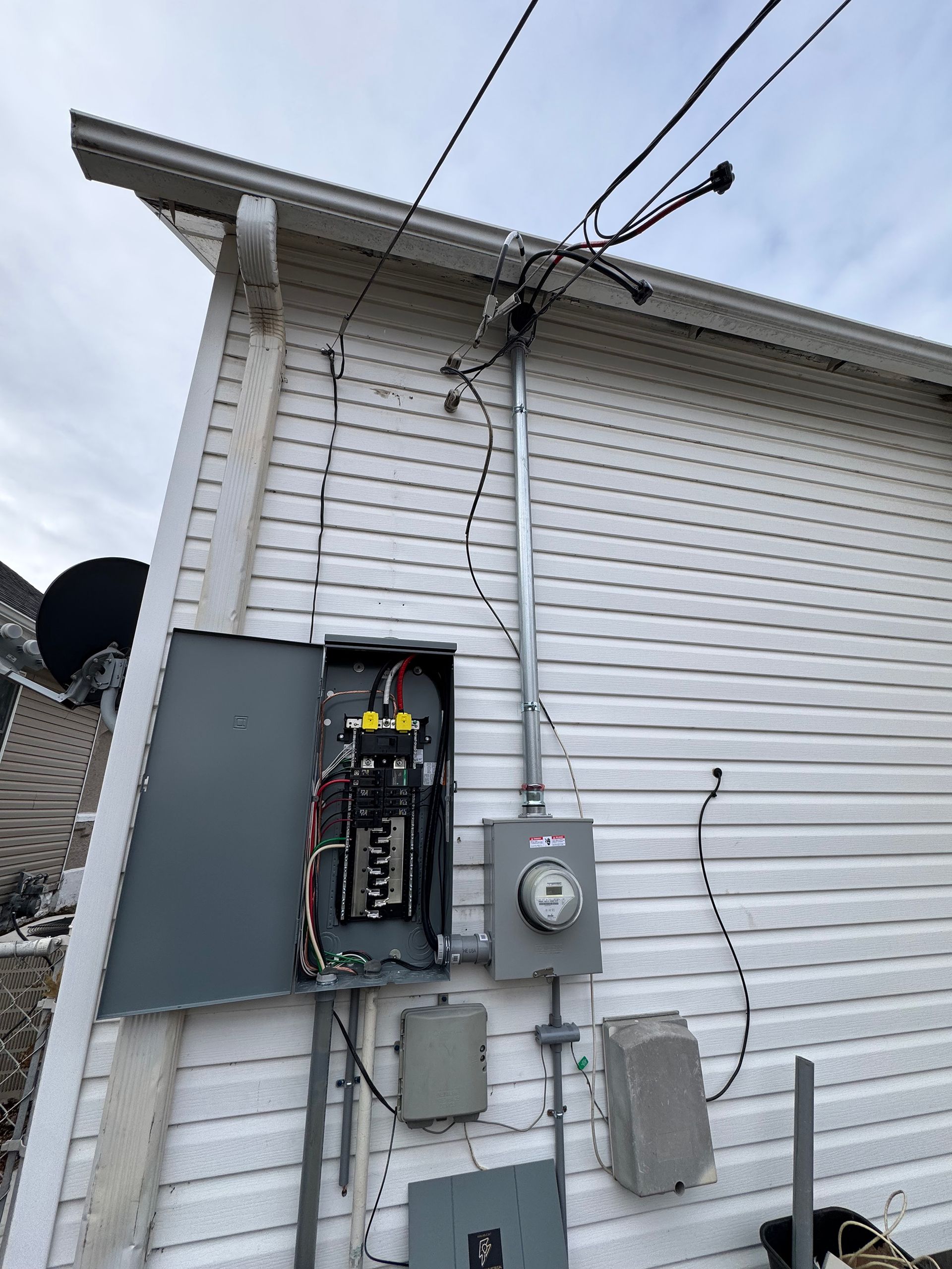 Exterior electrical panel with open panel door, meter, and wiring attached to a white-sided building.