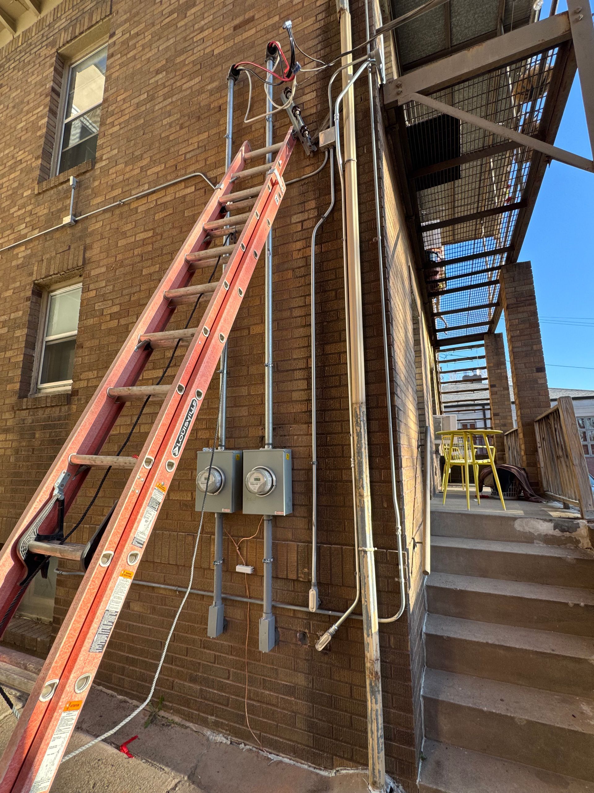 A red ladder leaning against a brick building, alongside electrical conduits and a fire escape.