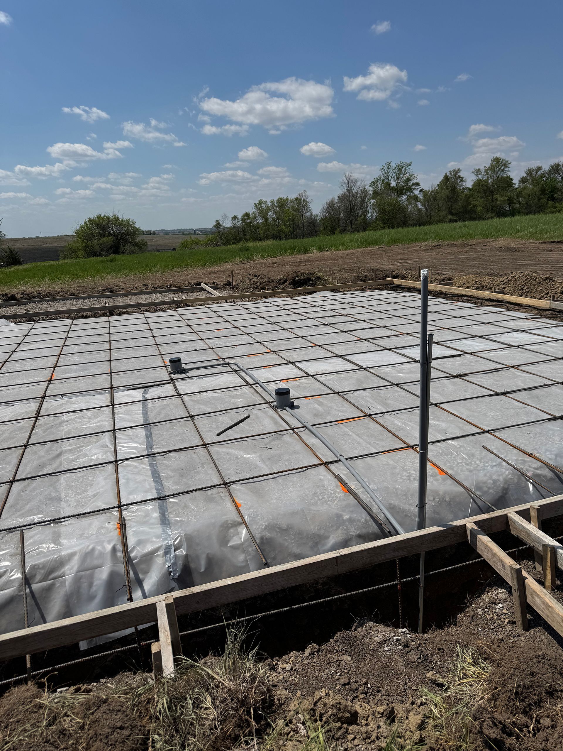 Construction site: concrete foundation form with rebar grid covered in plastic, against blue sky.