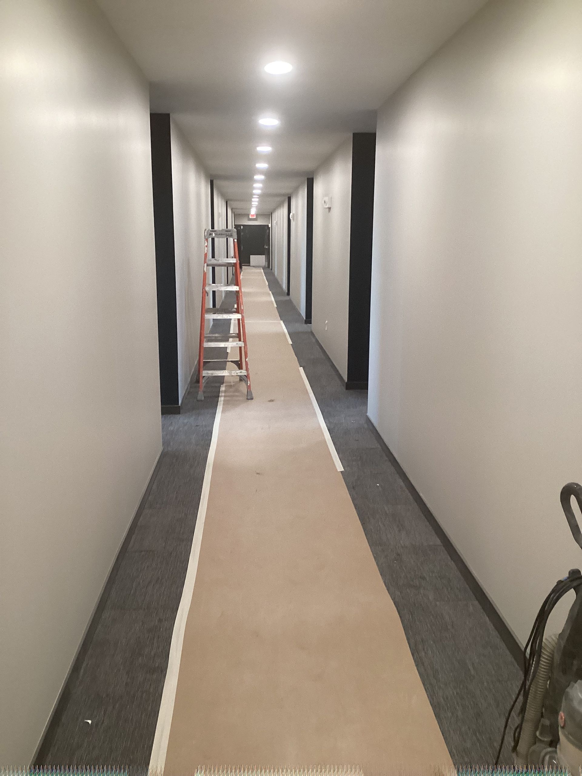 Long hallway with off-white walls, gray carpet, and a tan runner. A ladder stands mid-hall.