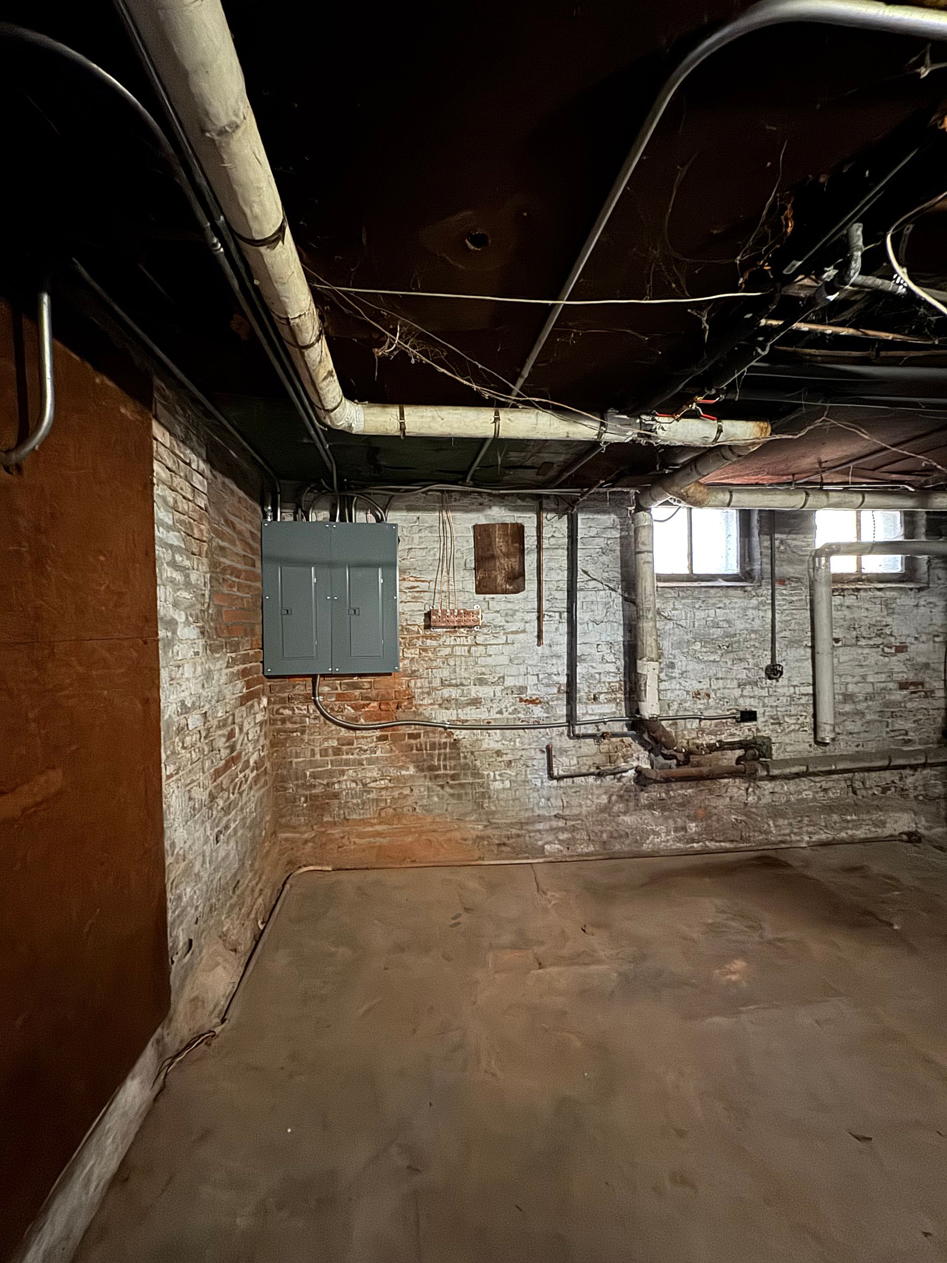 Basement with exposed brick walls, concrete floor, pipes, and electrical box.