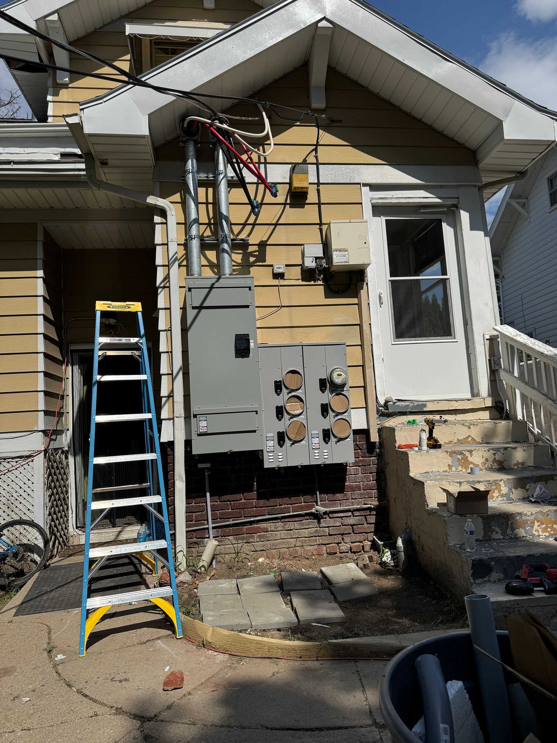 Ladder near electrical box on a building's exterior with wires. A door and stairs are visible.