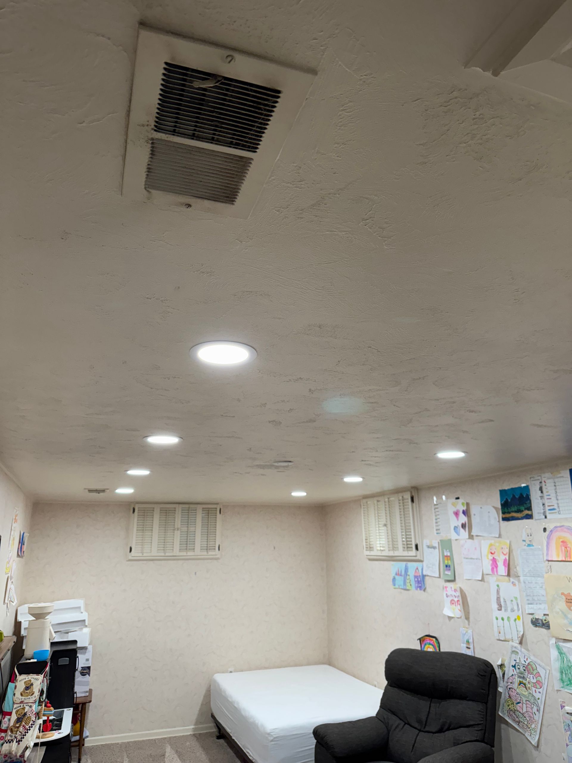 Basement interior with recessed lights, ventilation, bed, armchair, and artwork on the walls.