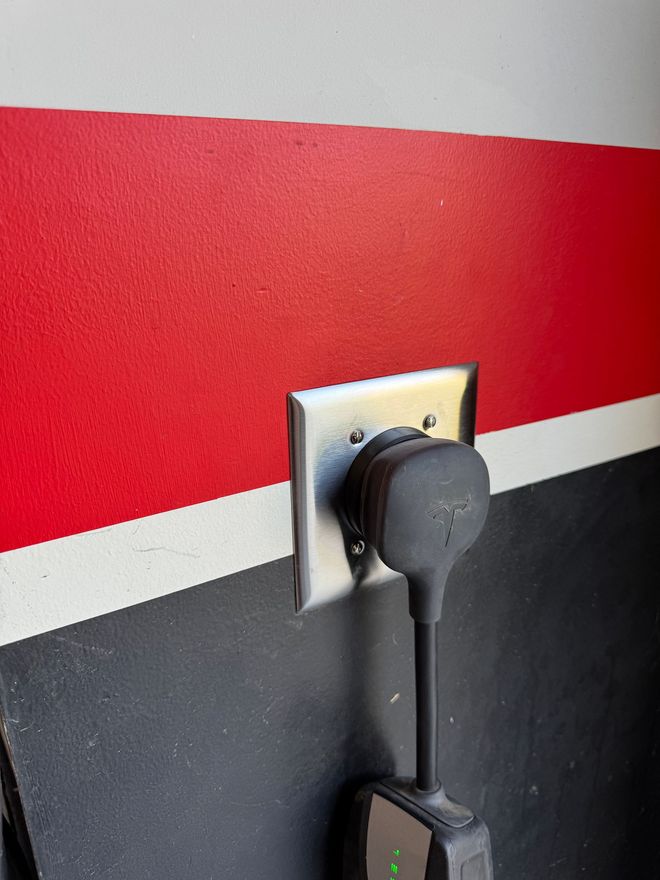 Black Tesla charger plugged into an outlet on a wall painted red, white, and black.