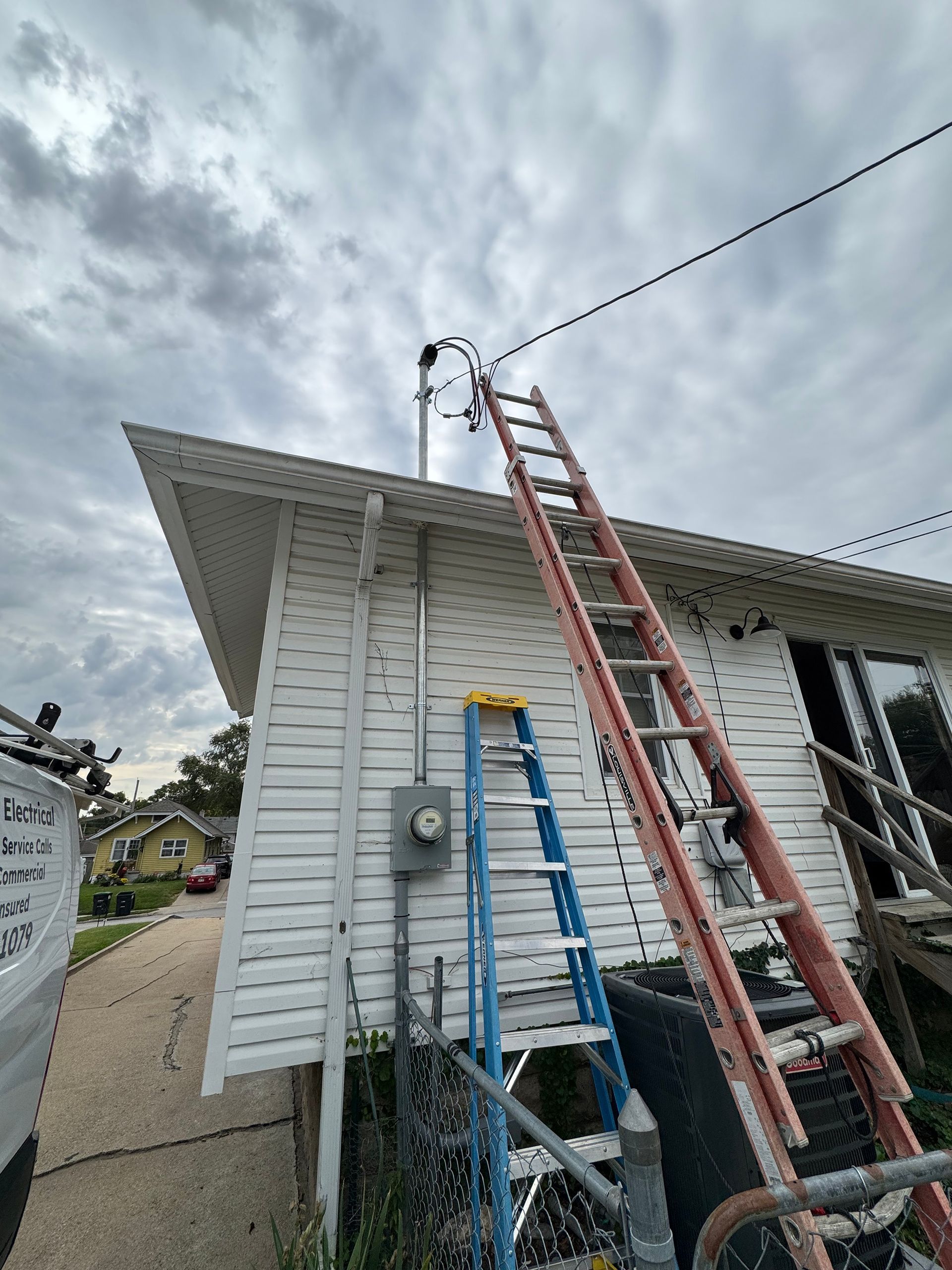 Electrical panel installation on a house exterior. Wires, ladder, and a door are visible.