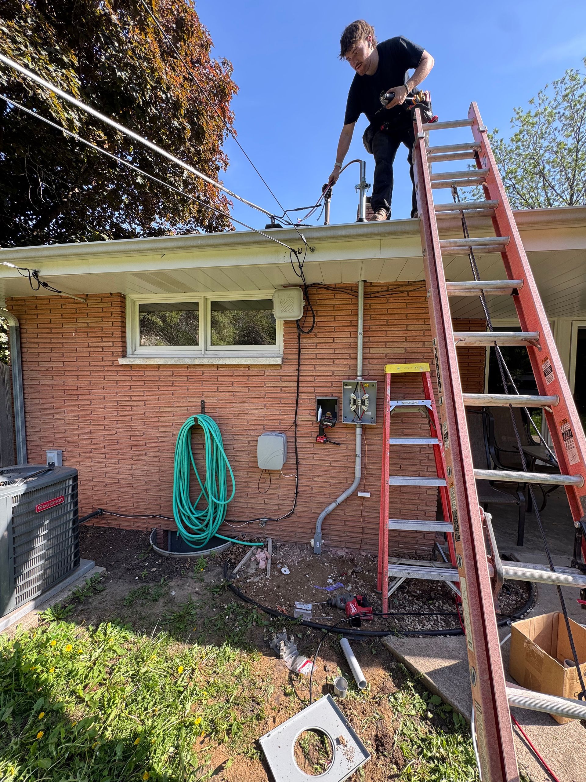 Man on ladder, working on electrical wiring near roof. Brick building, green hose, clear sky.