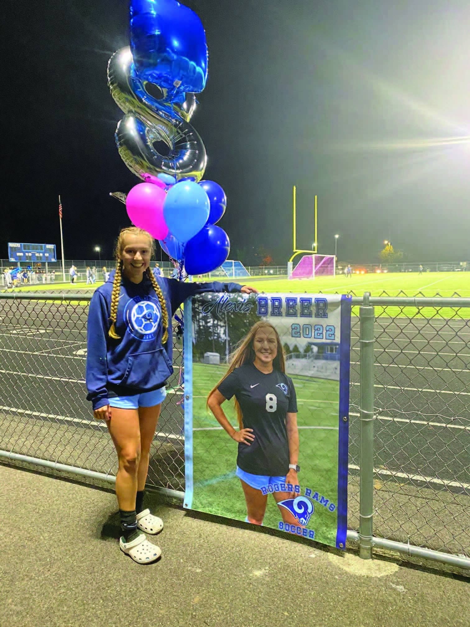 Teen girl stands by a fence with a poster of herself. She is holding balloons at a soccer field.