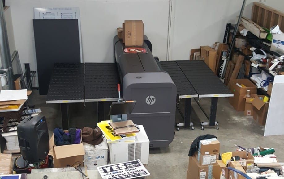 A large HP printer in a cluttered workshop; various boxes and items surround the machine.