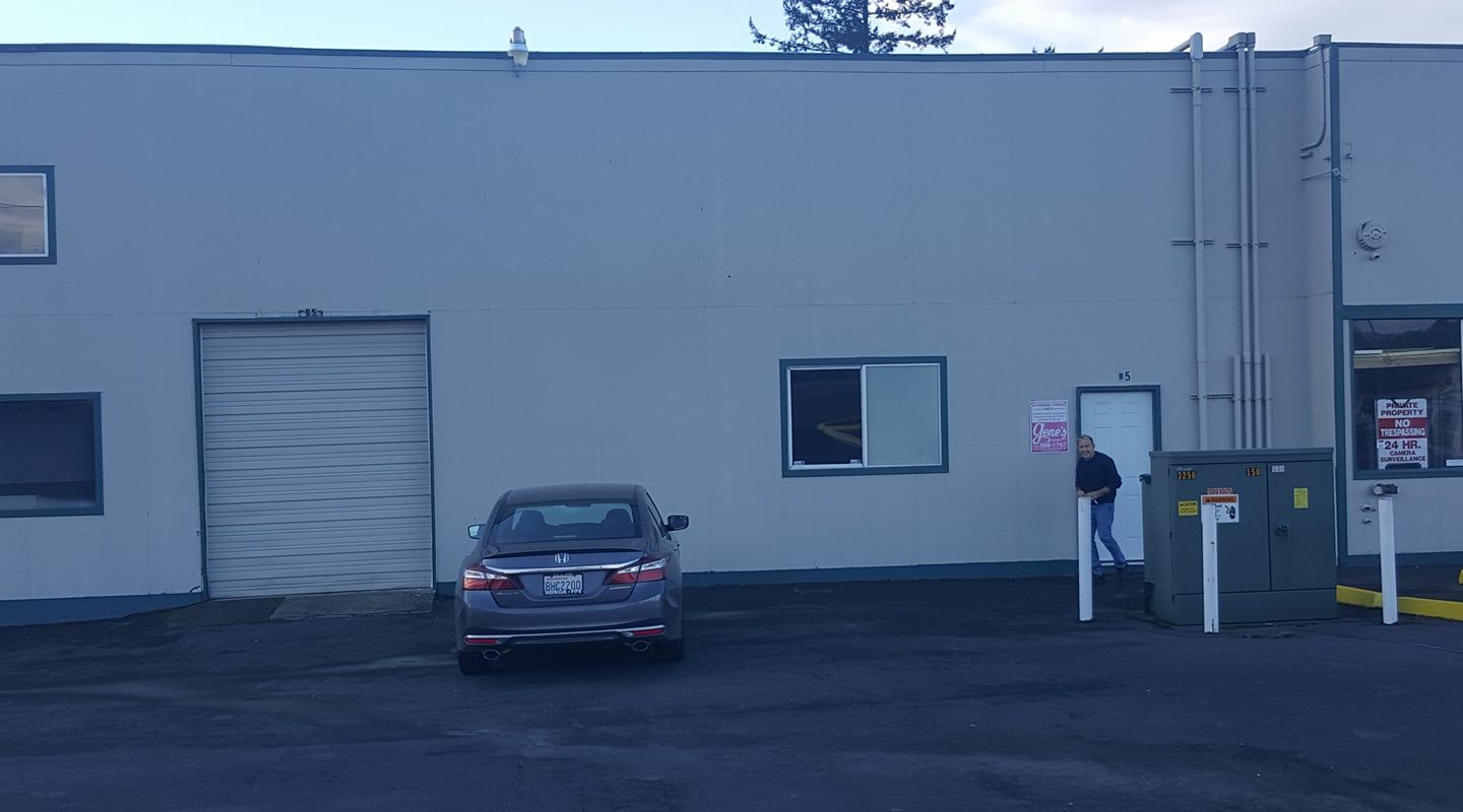 A gray car parked near a white building with a loading dock. A person exits a doorway holding a sign.