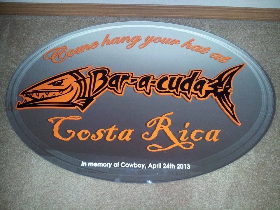 Oval sign for Bar-a-cuda bar in Costa Rica. Orange barracuda graphic and text on gray background. In memory of Cowboy.
