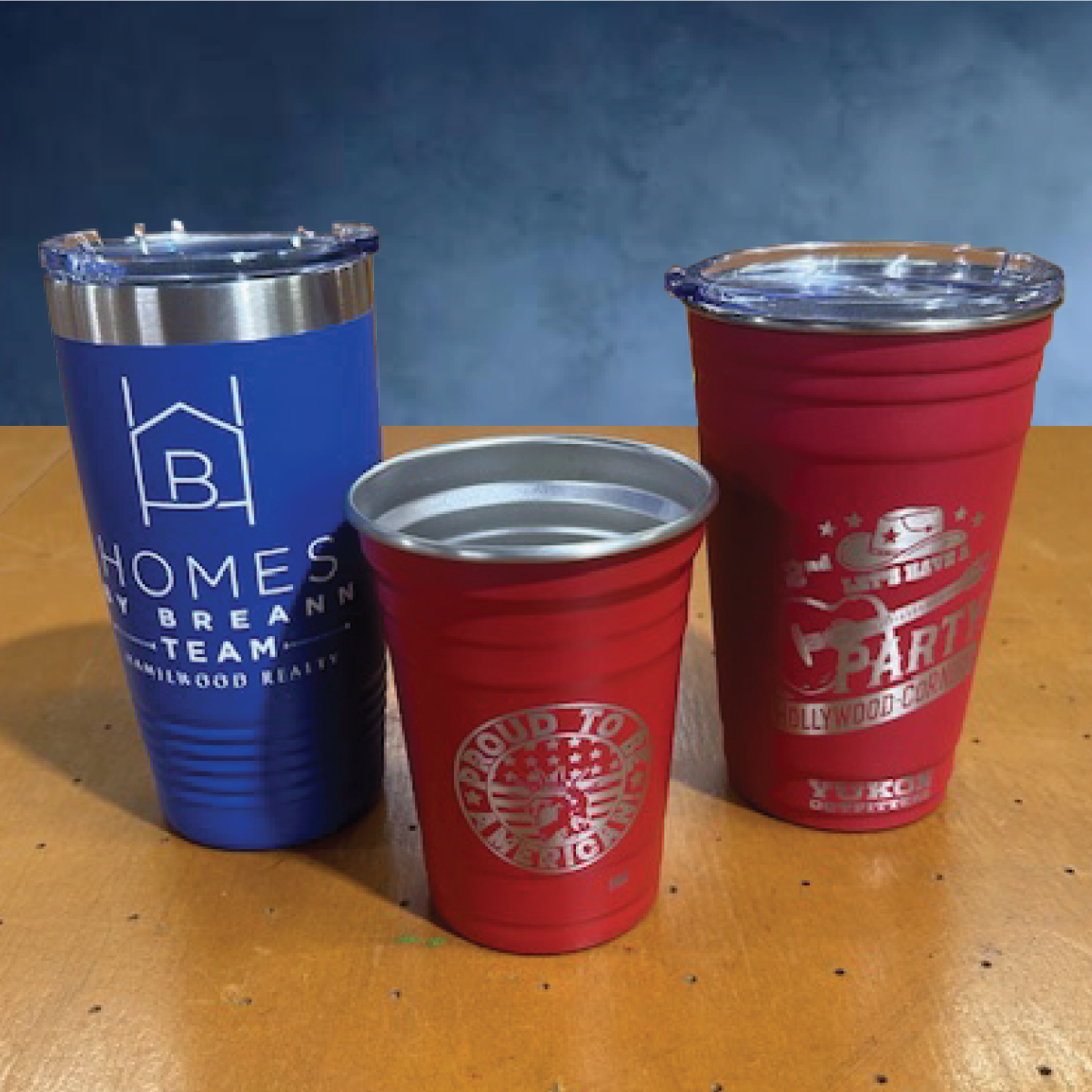 Three promotional metal tumblers on a wooden table: a blue one and two red ones with different logos and branding.