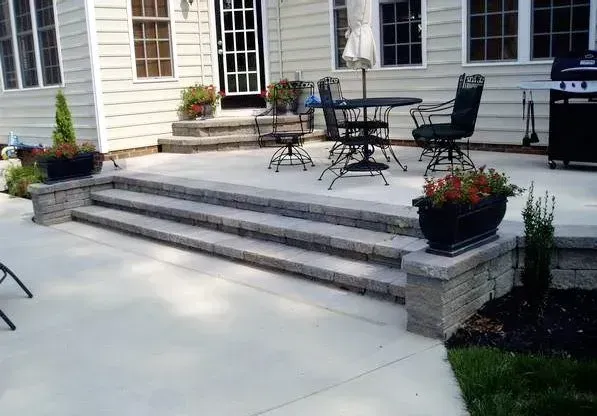 Stone steps lead to a patio with outdoor furniture, potted flowers, and a house.