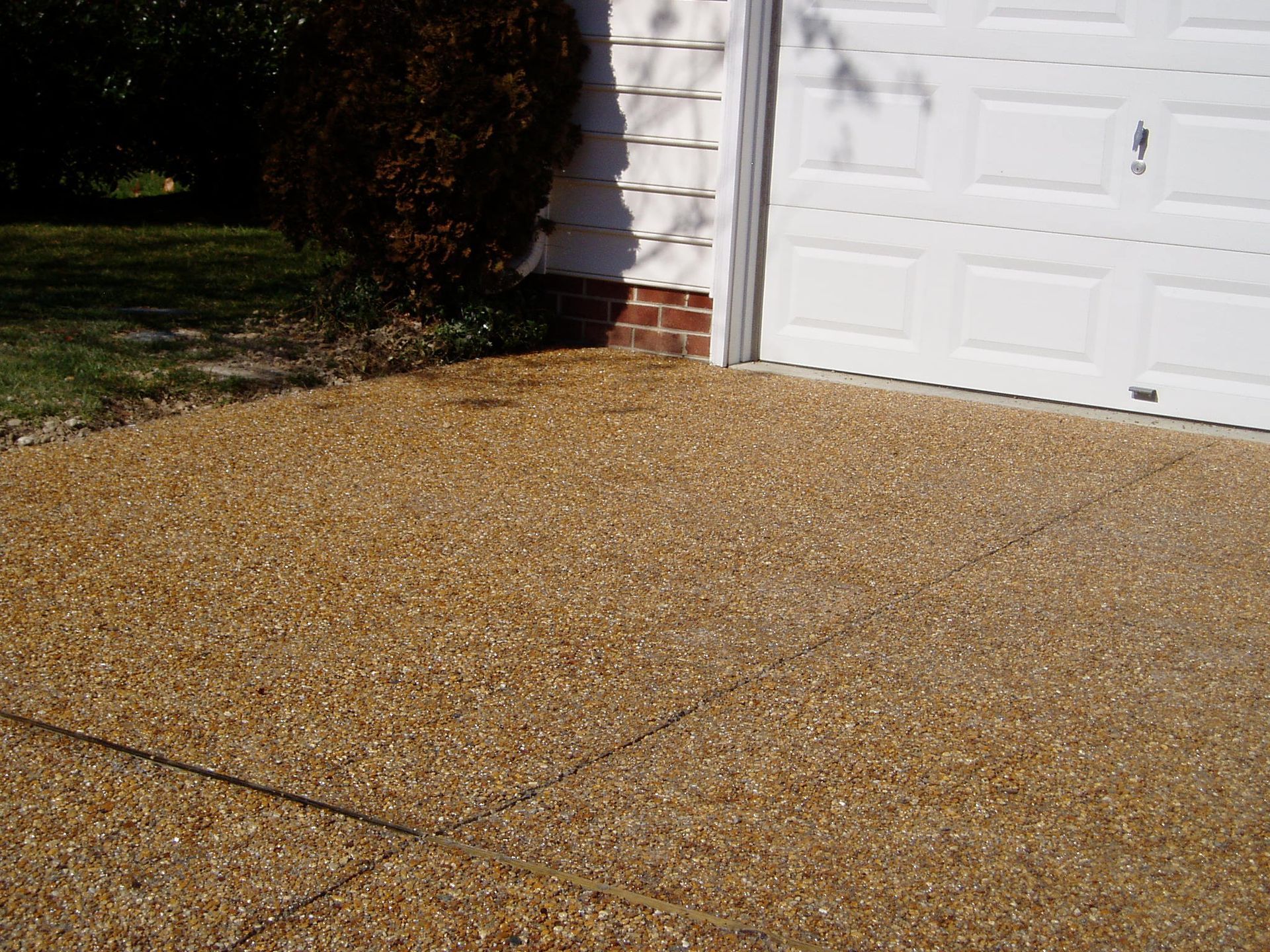 Tan aggregate driveway next to a white garage door, with a patch of green grass to the left.