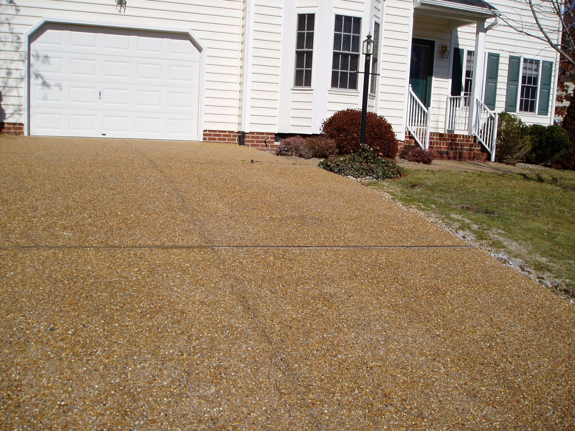 Tan pebble driveway leading to a white house with a garage door and small front yard with shrubs.