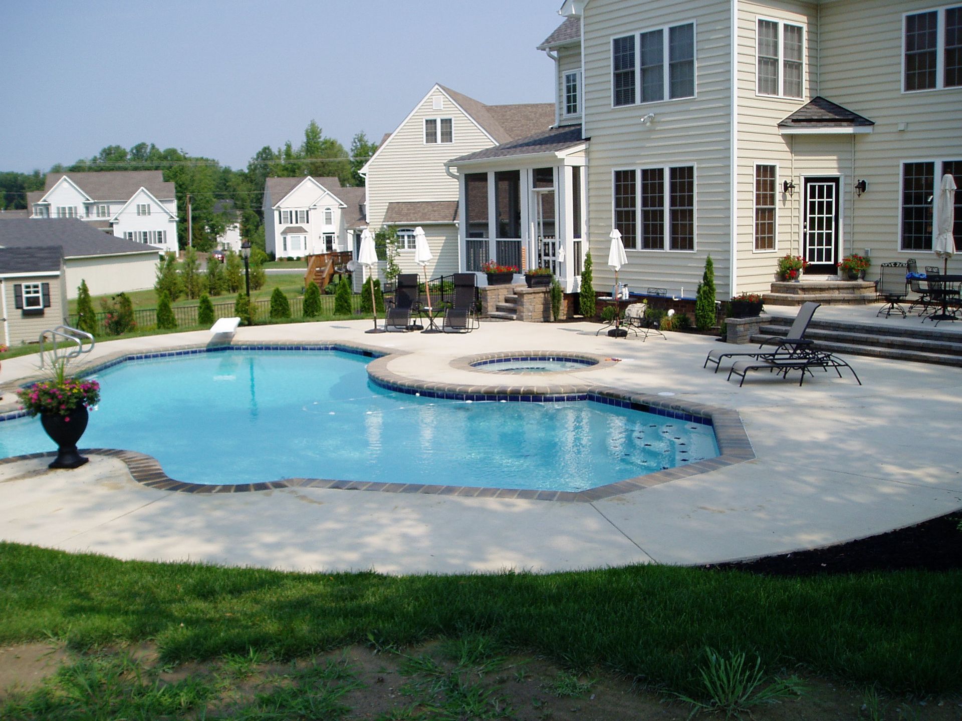 Backyard with pool, patio, and house. Blue pool, beige patio, multi-story house, and green lawn.