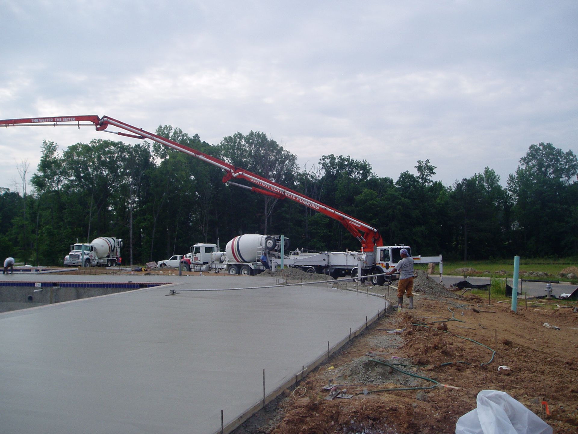 Concrete being poured from a boom pump onto a concrete pad at a construction site. Trucks, trees, and a worker are visible.