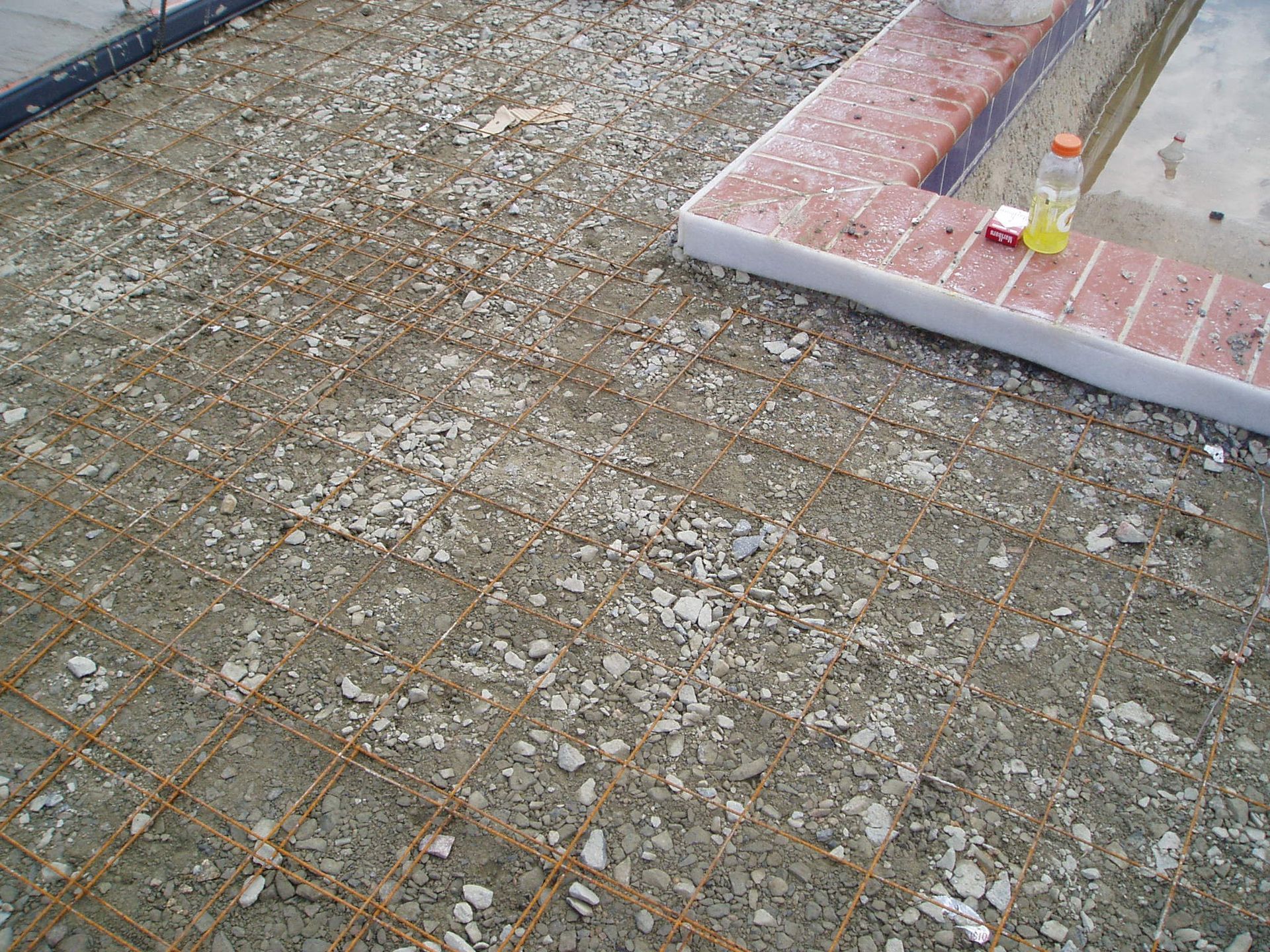 Concrete foundation with exposed rebar, gravel, and part of a brick pool border.
