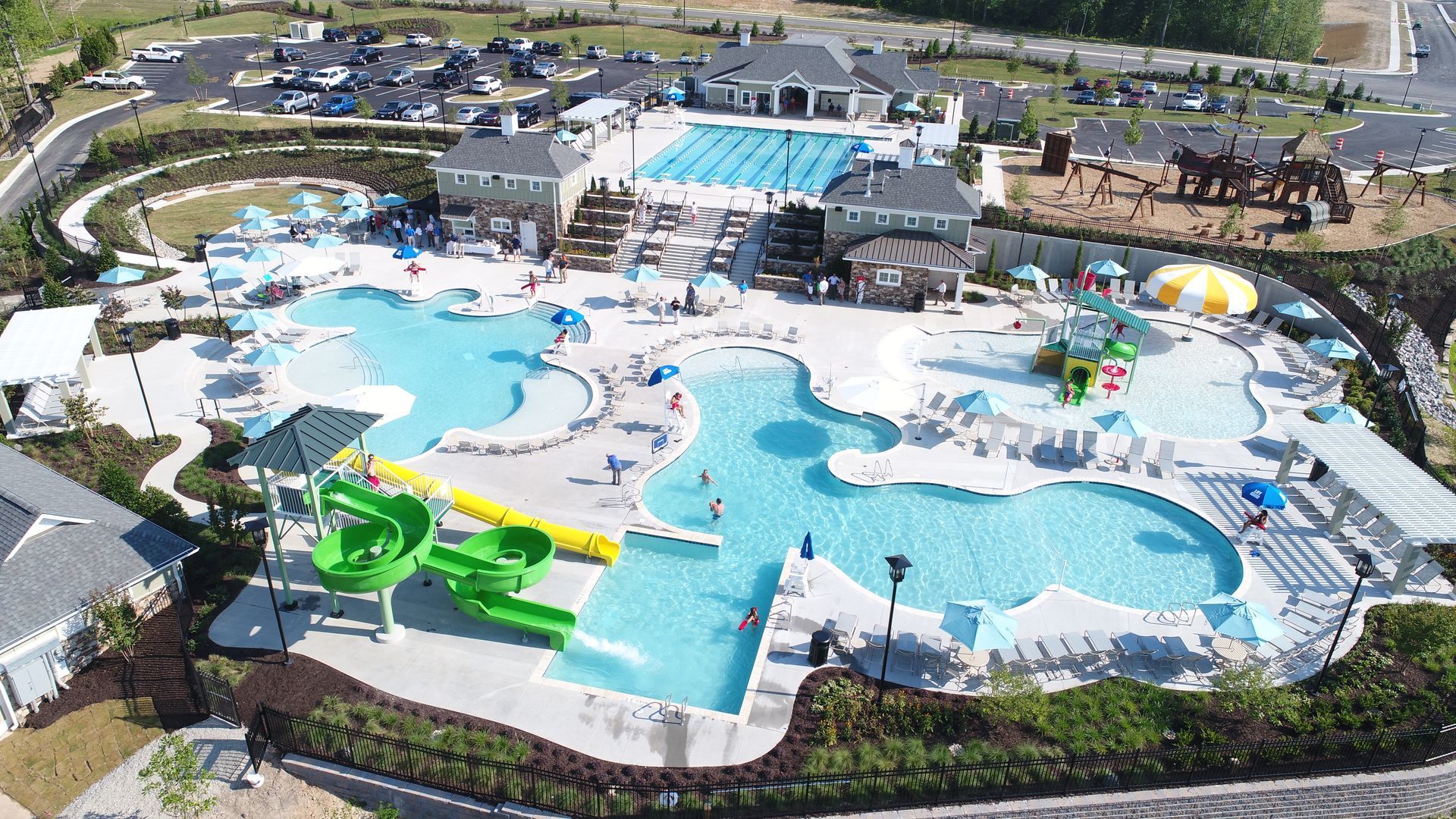 Aerial view of a large community pool complex with several connected pools, water slides, and buildings; blue water, green grass.