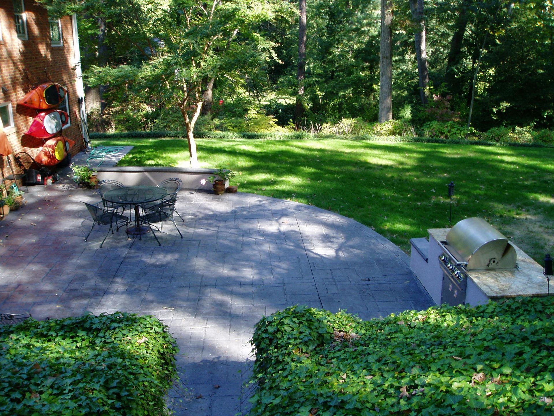 Backyard patio with a grill, table, and green lawn surrounded by trees.