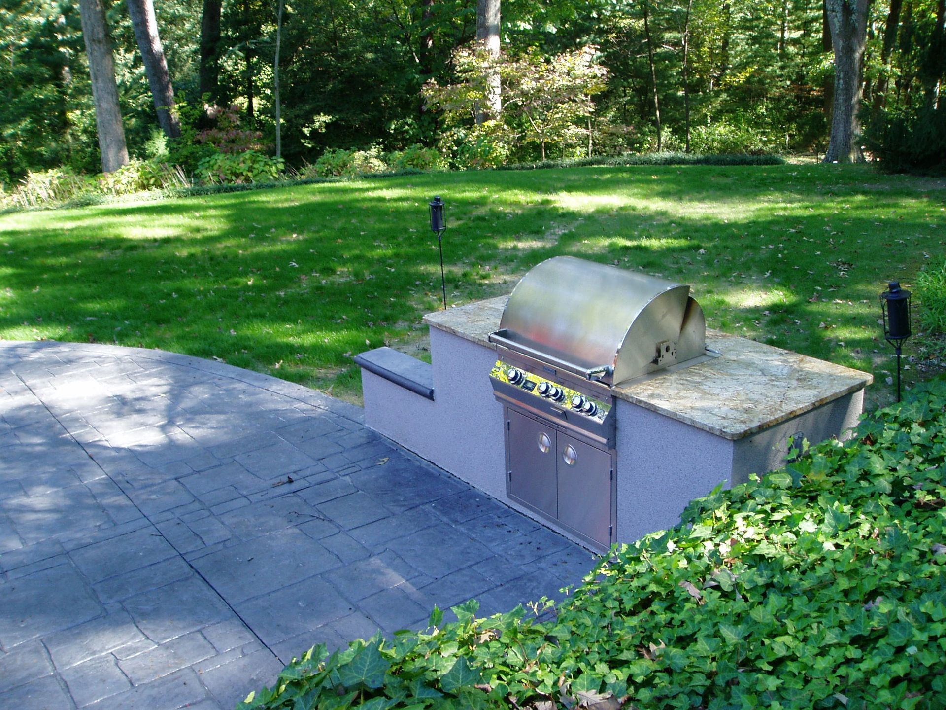 Outdoor kitchen with built-in grill on a gray stone structure, surrounded by green lawn and trees.