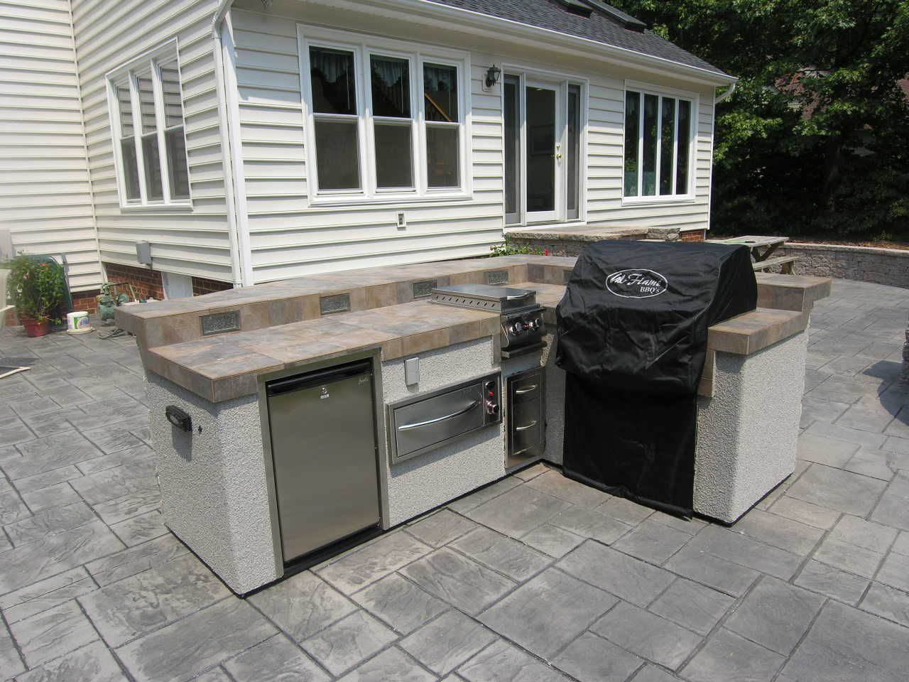 Outdoor kitchen with concrete countertop and built-in grill, refrigerator, and side burner on a patio.