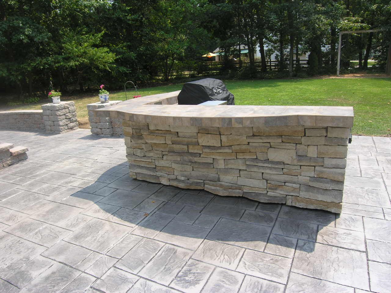 Stone outdoor kitchen with built-in grill and concrete patio in a backyard setting.