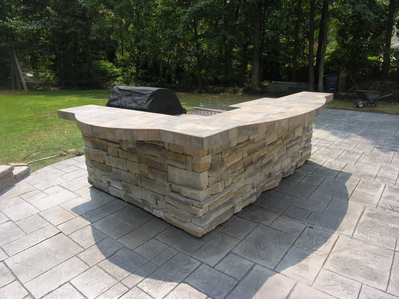 Stone outdoor kitchen on stamped concrete patio. Grill on countertop, surrounded by green grass and trees.