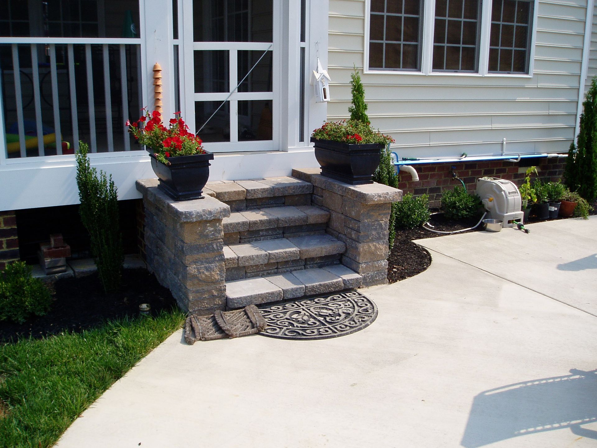 Stone steps leading to a white door, flanked by flower pots. Concrete walkway and green grass.