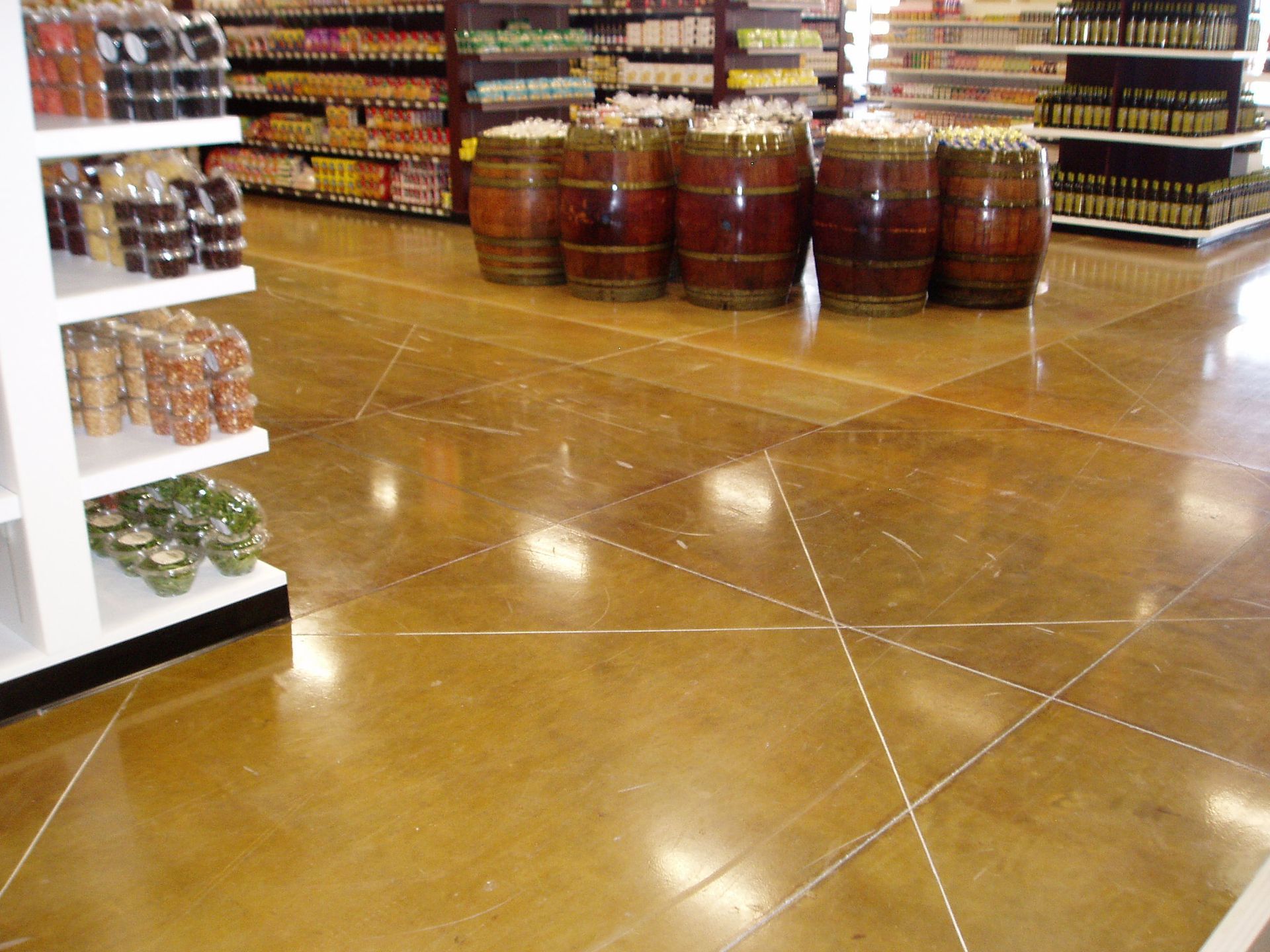 Polished concrete floor in a grocery store aisle, reflecting light. Wooden barrels and shelves of products are visible.